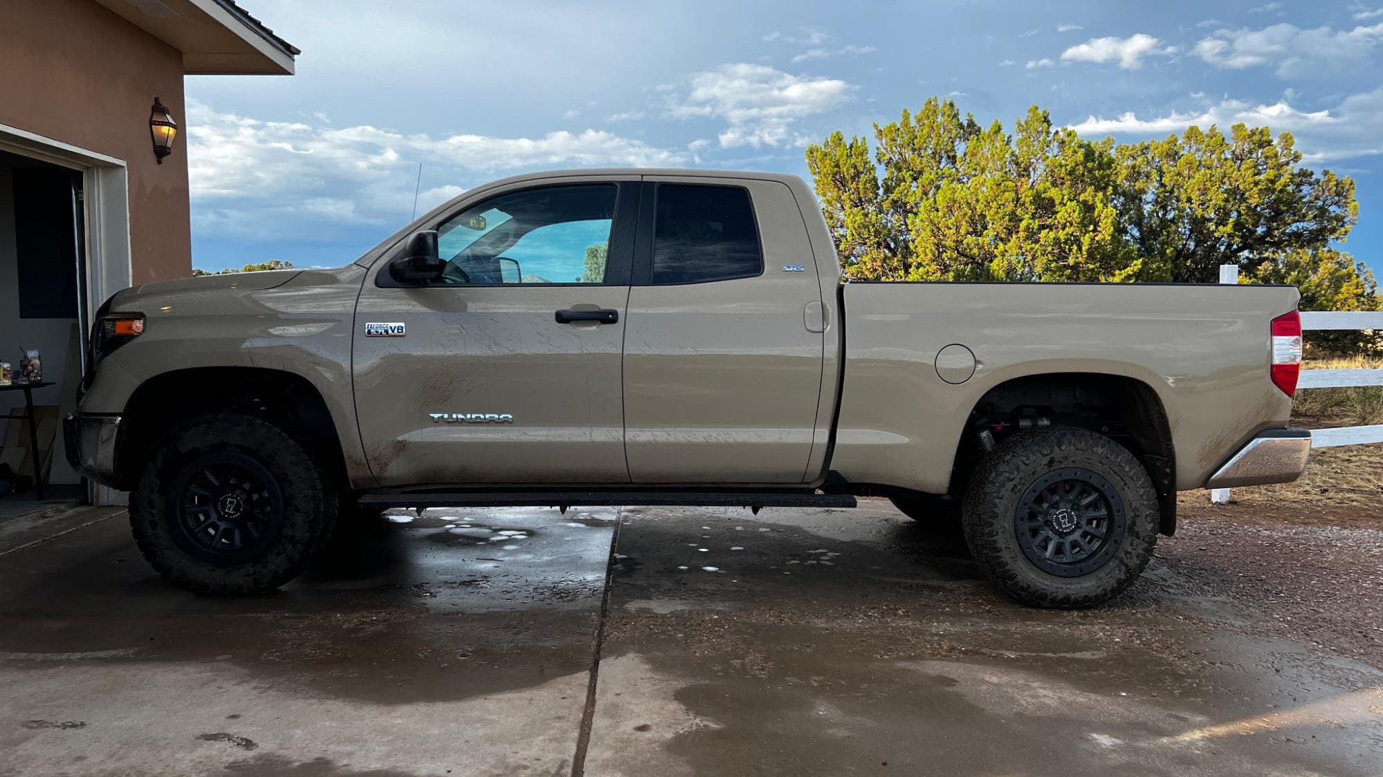 Tan Toyota Tundra with lifted suspension and large off-road tires parked in a driveway, mud splattered along the doors and rocker panels.