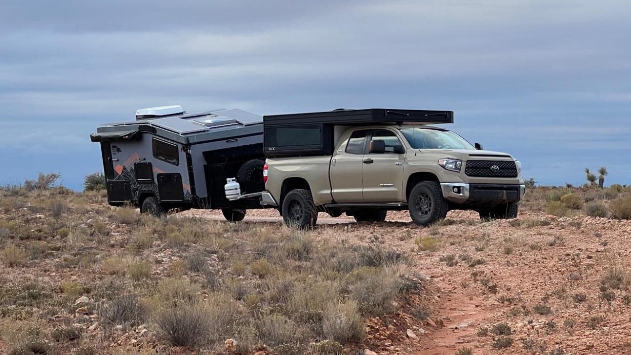 Tan Toyota Tundra towing an off-road camper trailer along a dirt road under an overcast sky.