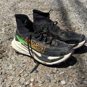 Pair of well-worn black Hoka Tecton X 4 trail running shoes with sock-like high ankle collars resting on gravel ground.