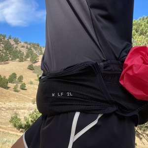Side profile of a runner wearing a black trail running waist belt with a bright pink jacket stashed in the rear storage loops on a hillside trail.