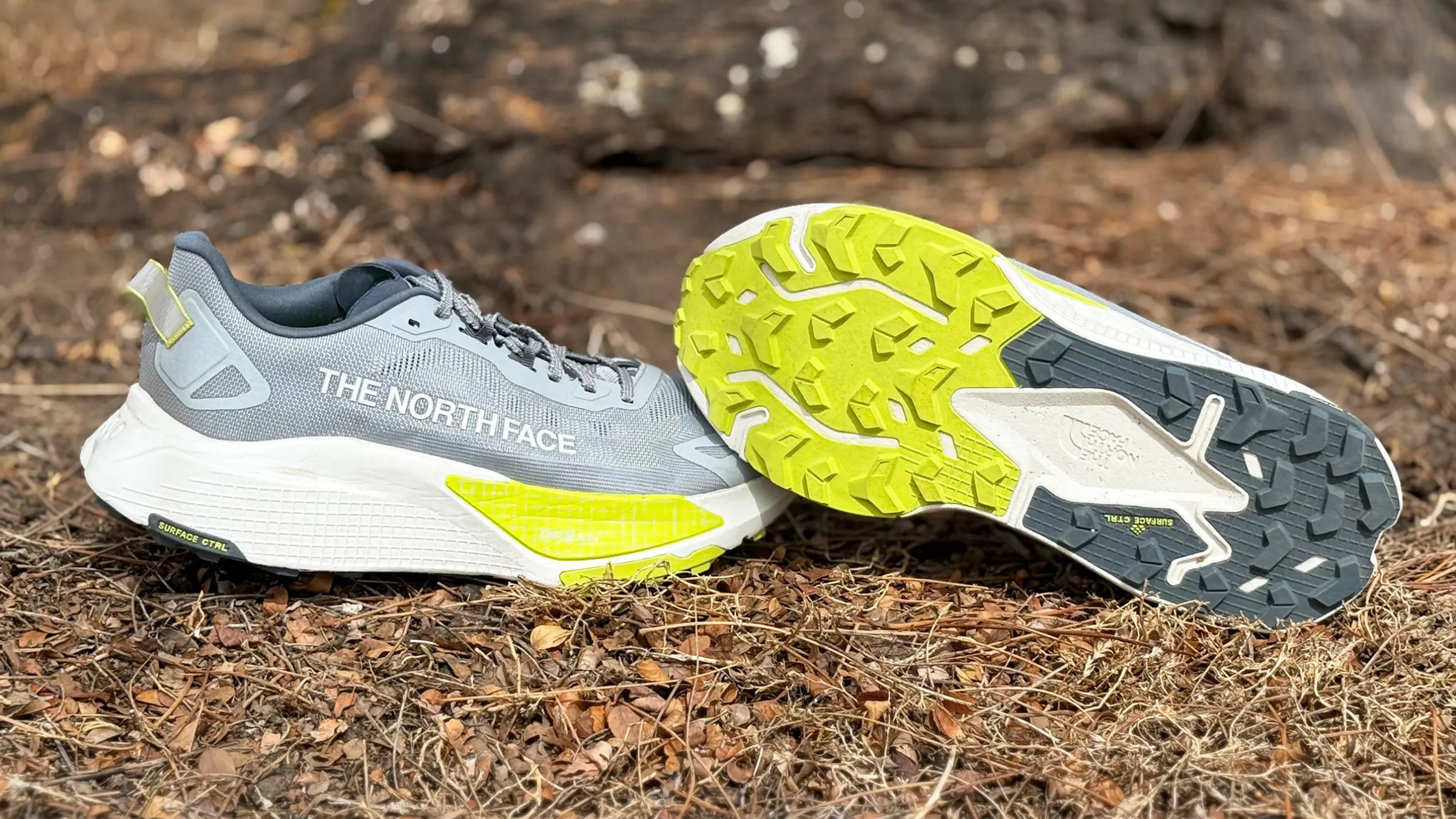 Grey and neon-yellow The North Face trail running shoes resting on forest soil, highlighting the grippy lugged outsole designed for off-road traction.