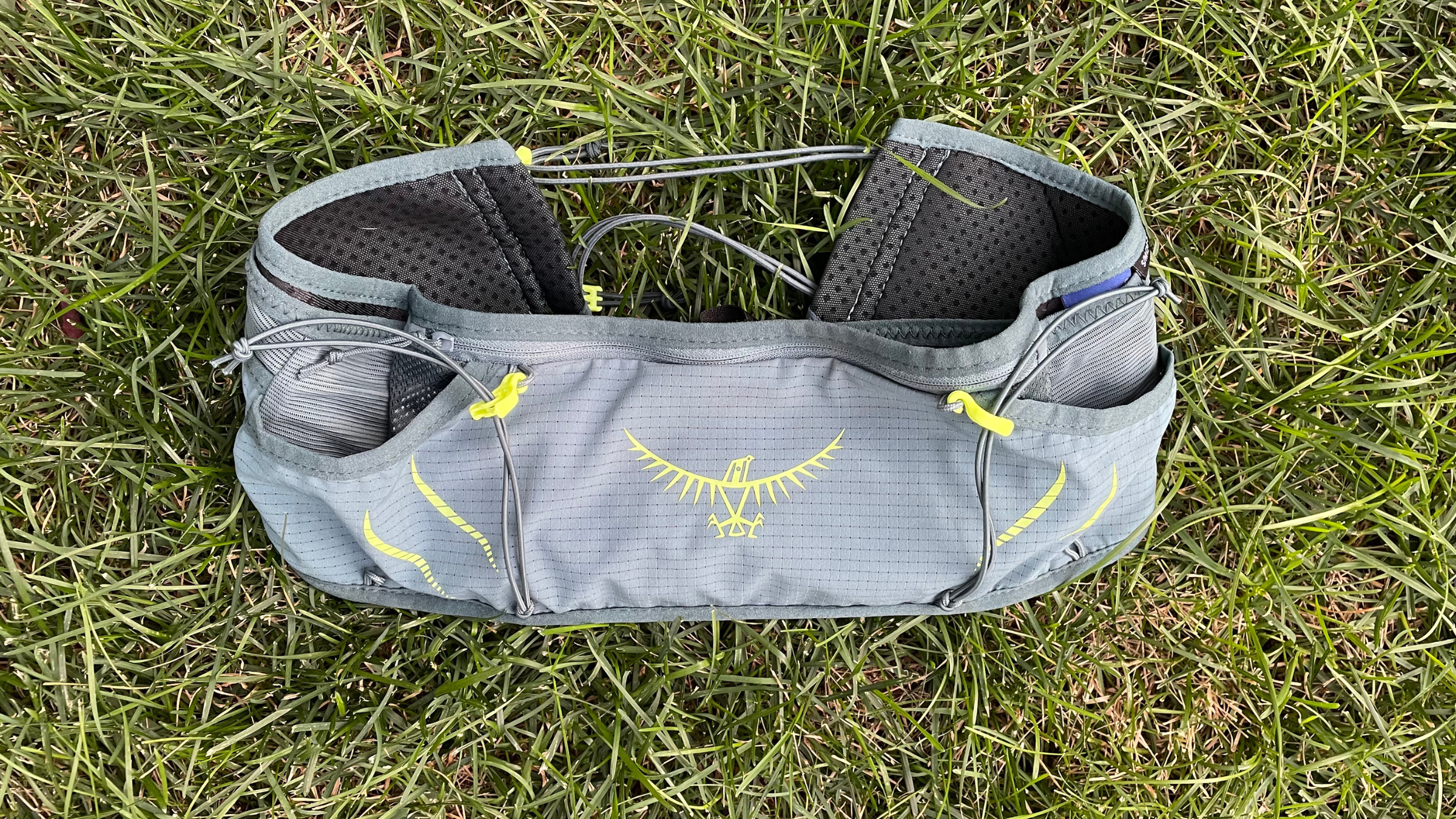 Grey Osprey trail running waist belt with high-visibility yellow cinch cords and logo lying flat on grass.