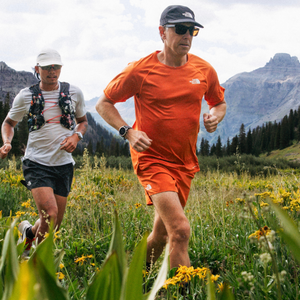 Writer Cory Smith wearing a bright orange running shirt and shorts runs down a mountain dirt trail filled with yellow wildflowers.