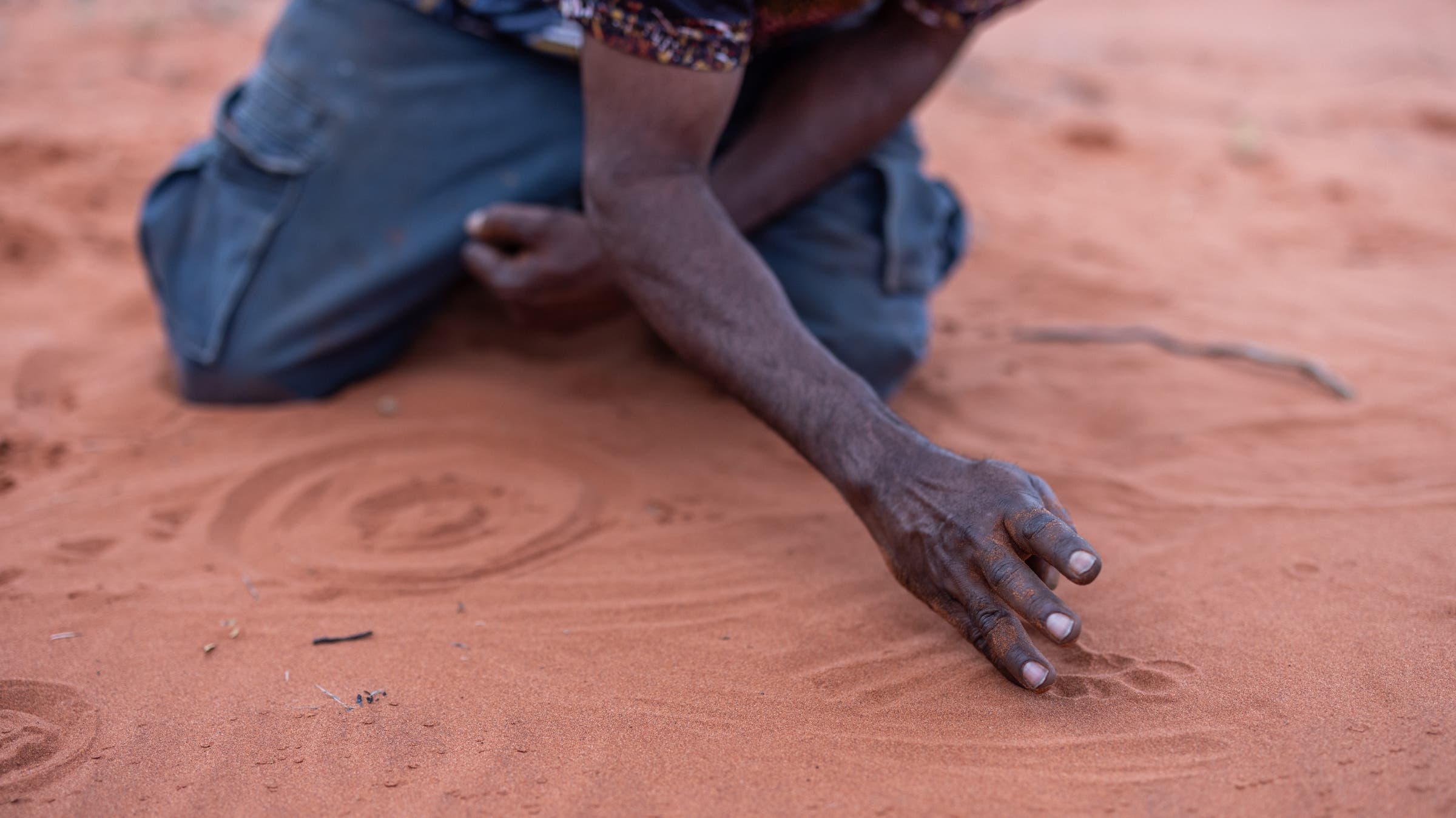 A man crouches down, drawing shapes in red sand.