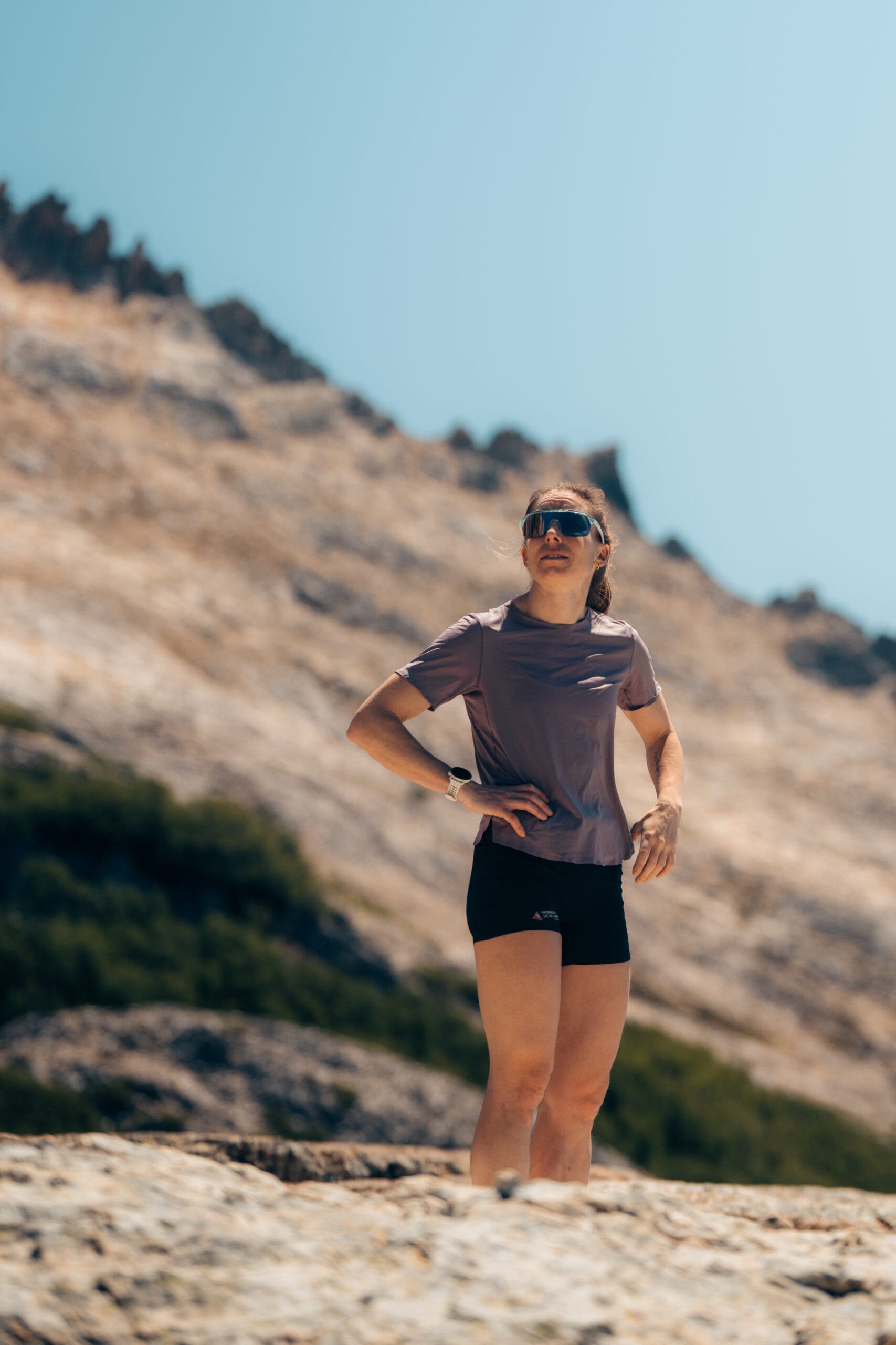 kalie mccrystal stands in front of a mountain wearing a purple shirt, black shorts, with her hands on her hips