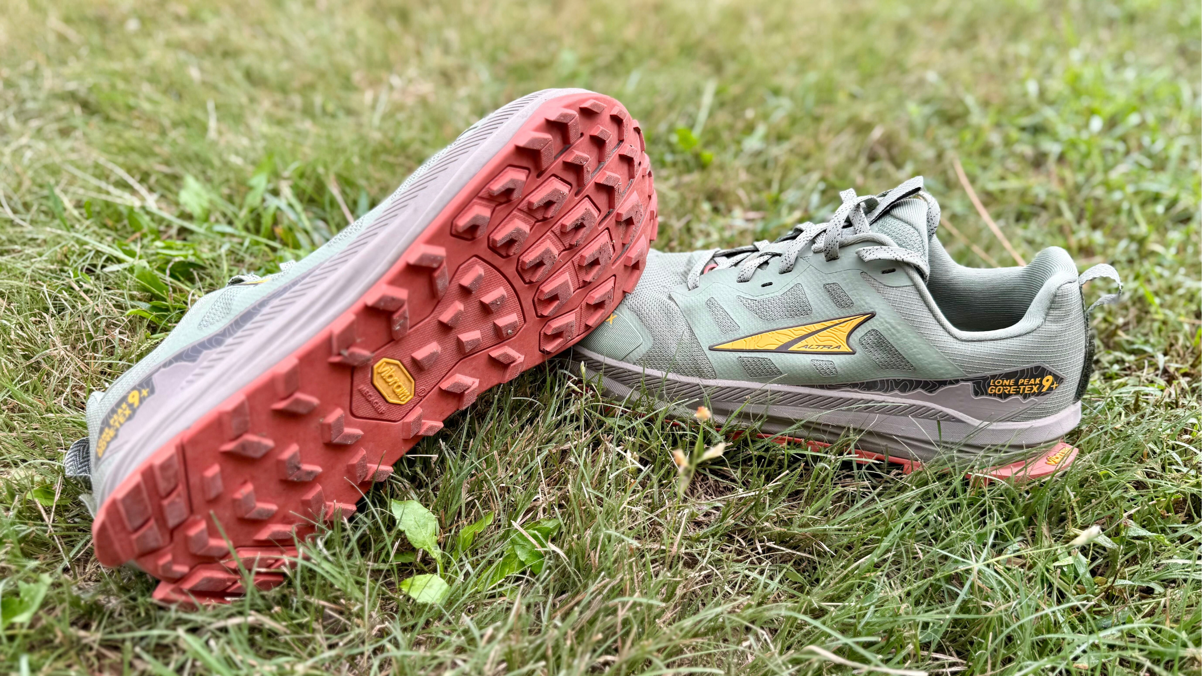 Pair of sage green Altra Lone Peak 9+ GTX trail hiking shoes resting on green grass, showing the red Vibram Megagrip lugged sole and Gore-Tex side badging.