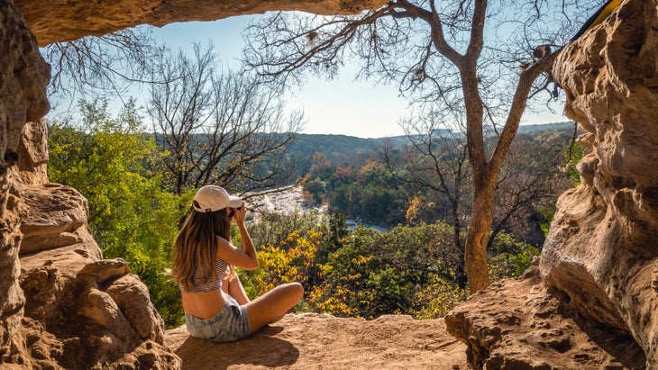 Woman sitting at the edge of a sunlit cave overlooking a river.