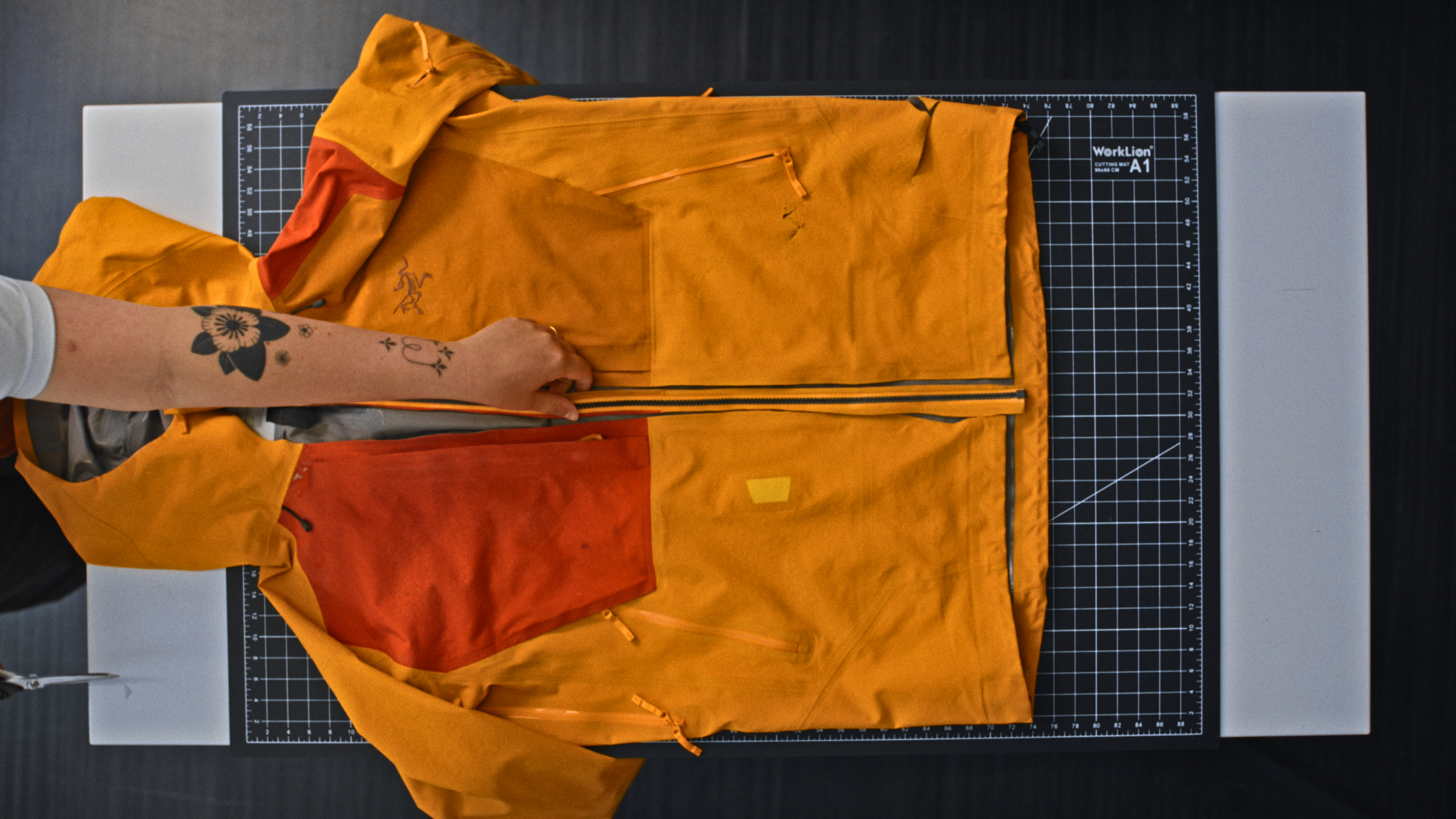 A top-down shot of an orange two-tone Sperro SV jacket laid flat on a grid cutting mat while a designer inspects the central zipper.