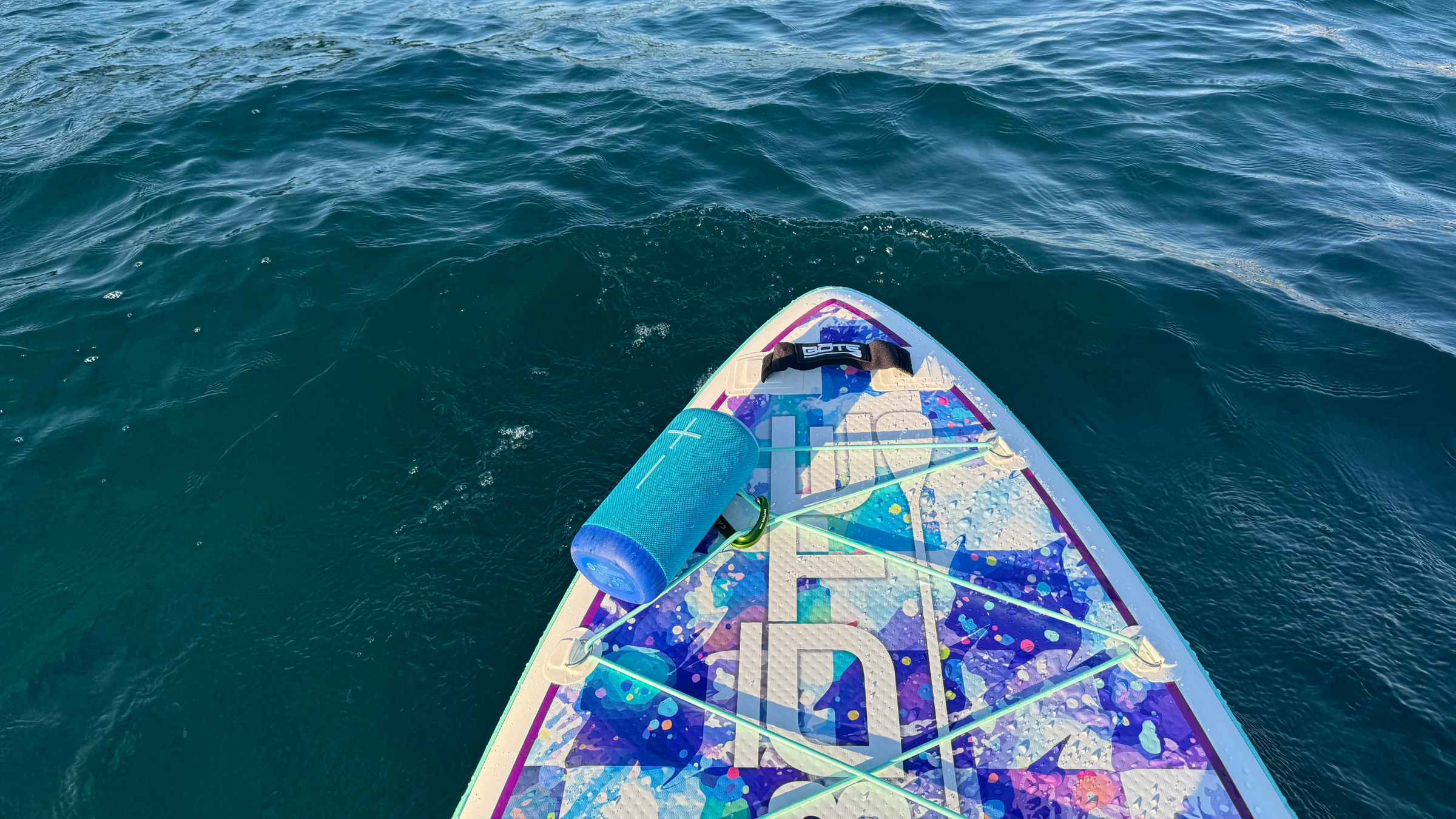 The bow of a colorful BOTE paddleboard floats on choppy blue water with a Bluetooth speaker secured beneath the front bungee tie-downs.