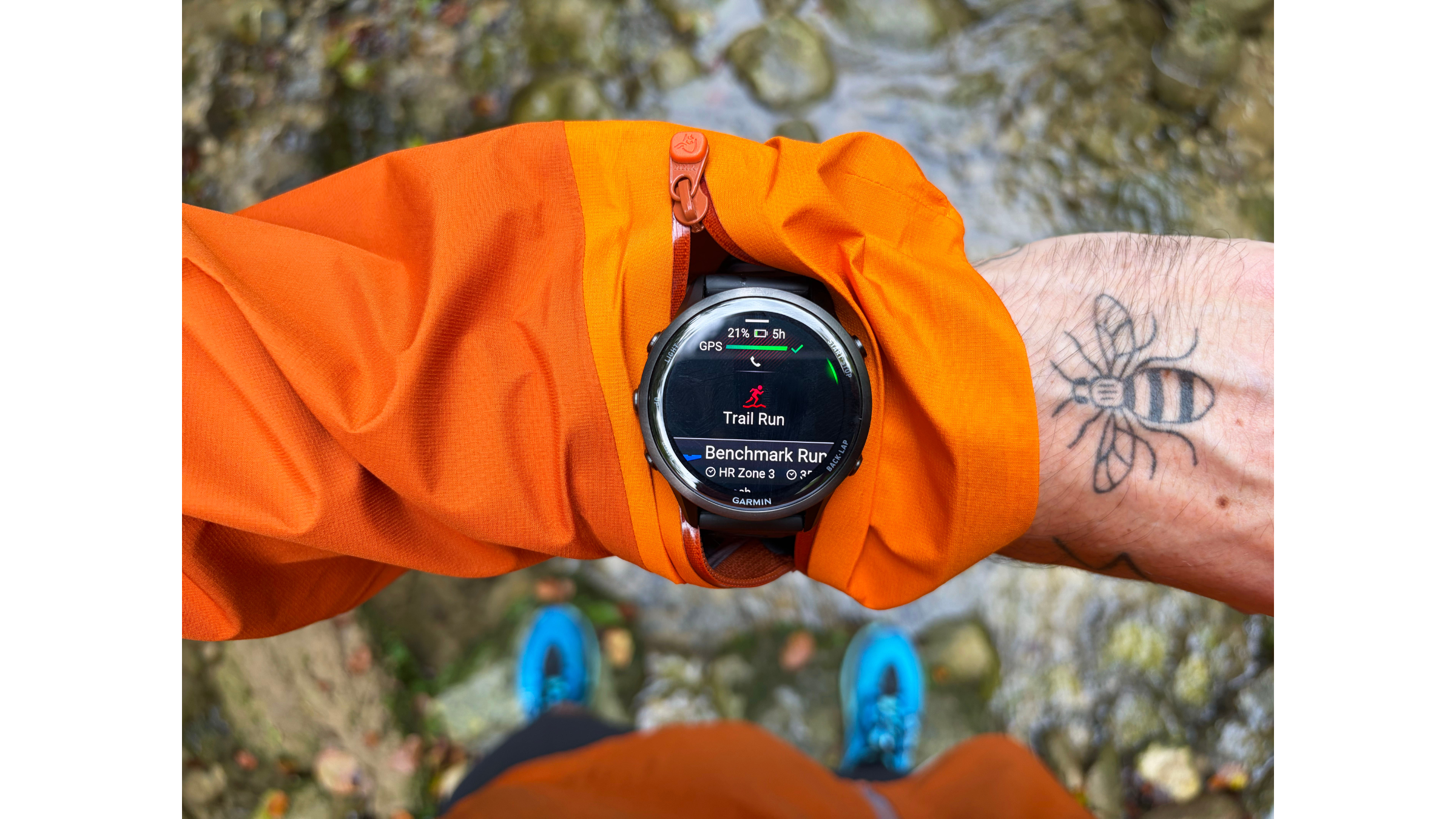 An overhead view focusing on a runner's wrist with a bee tattoo, wearing an orange Norrøna trail running jacket with a specialized zippered opening to view a Garmin GPS watch.