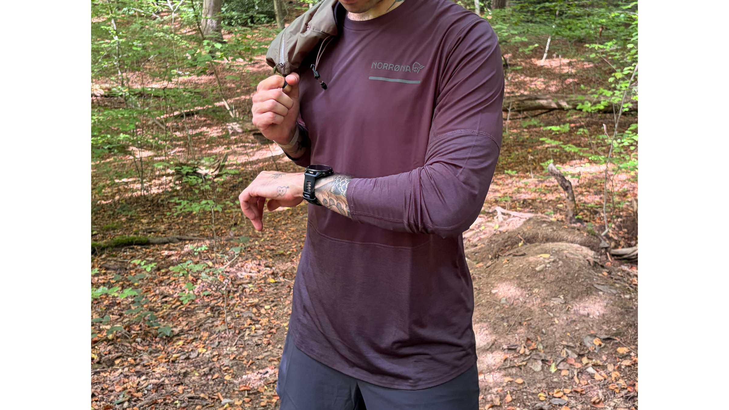 A close-up view of a runner in a maroon Norrøna trail running long-sleeve shirt, checking his watch on a forest path.