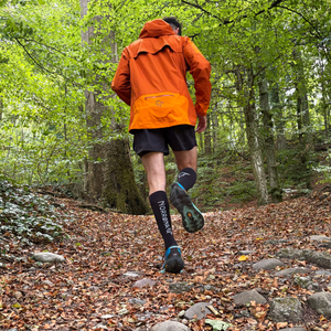 Rear view of a person wearing an orange Norrøna trail running jacket and branded socks, running uphill on a steep, leaf-covered forest trail.
