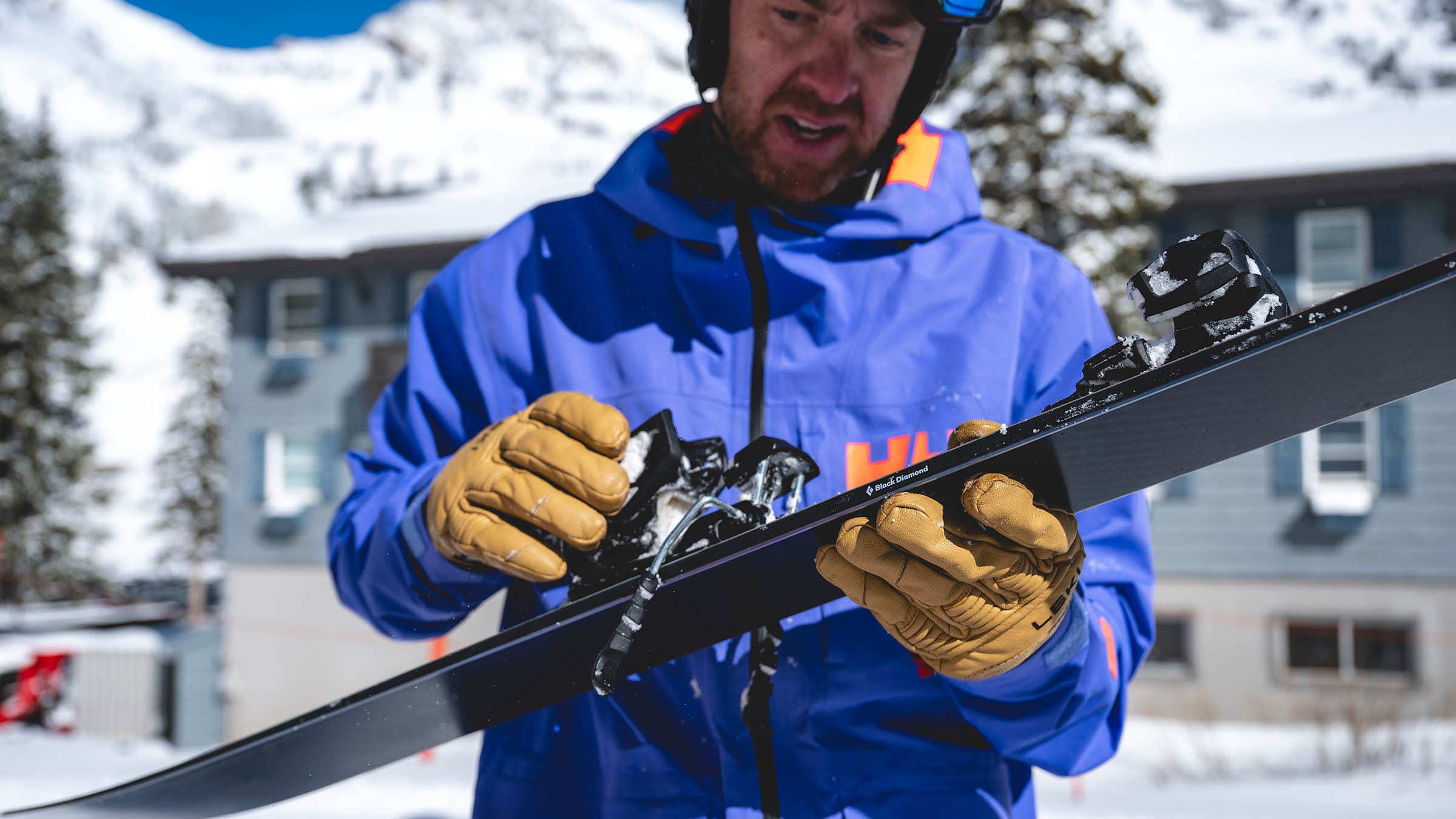 Skier in purple jacket adjusting ski bindings with leather gloves at ski resort base area