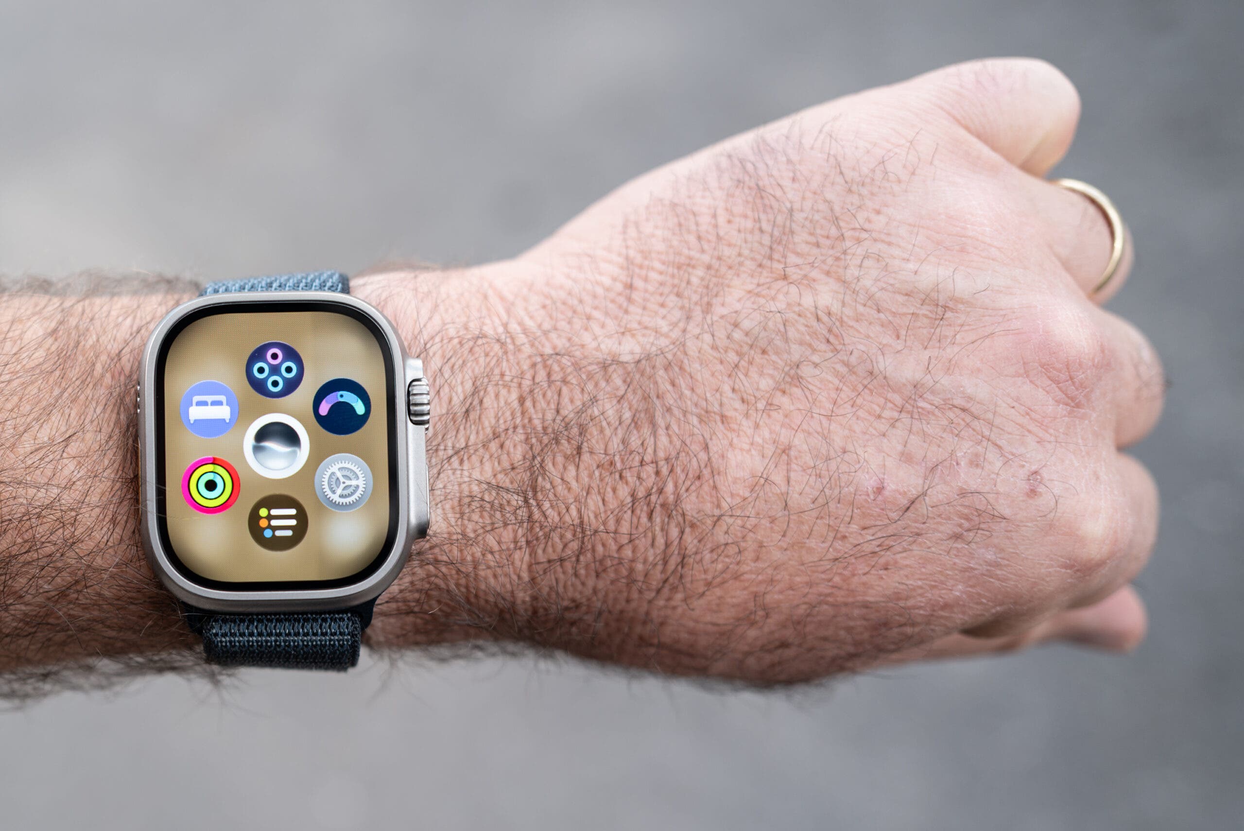 Apple Watch Ultra 4 with a titanium case on a hairy wrist, its screen showing a colorful grid of app icons, held in front of a gray background with a dark woven band.