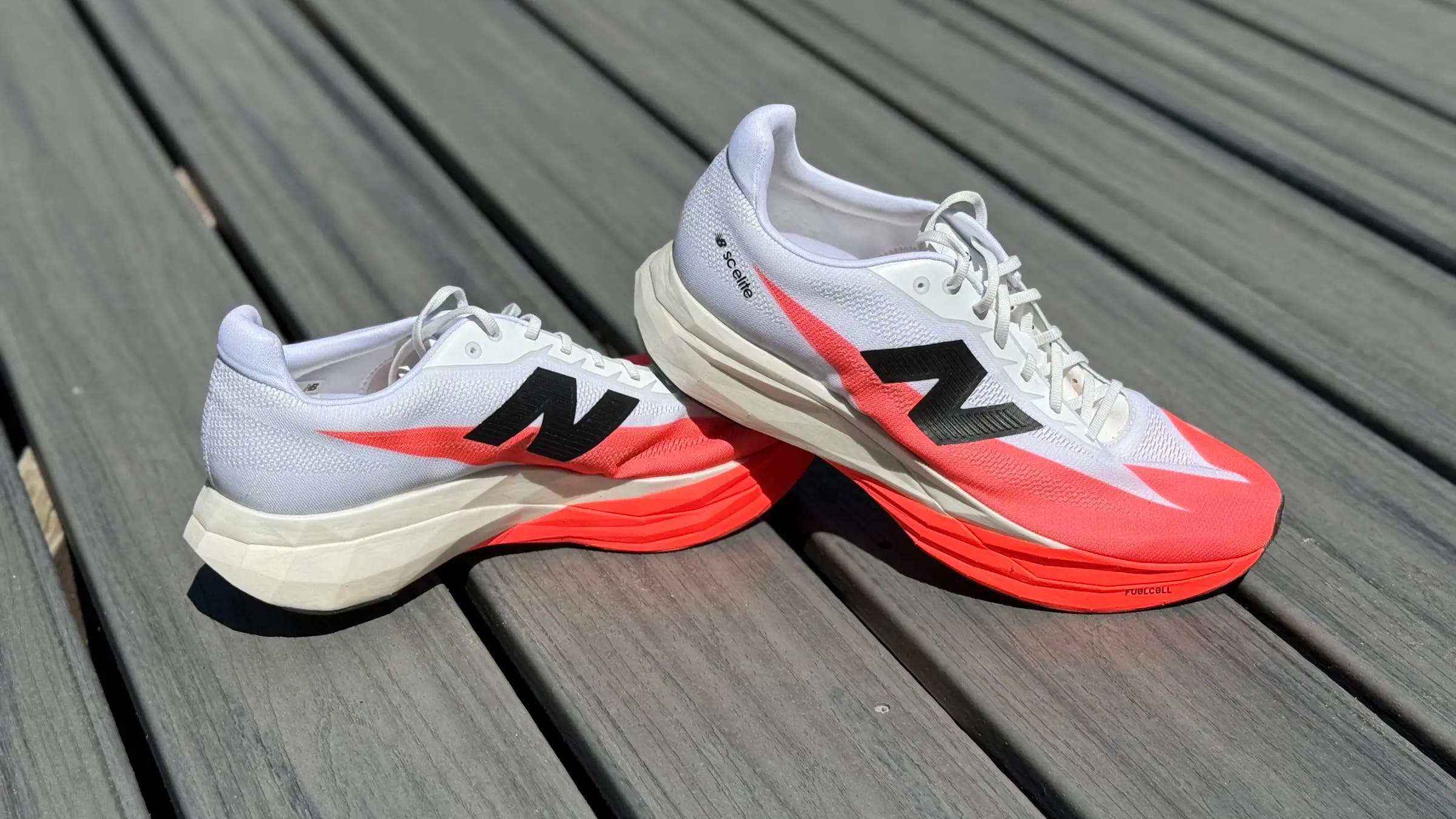 Pair of white and red-orange New Balance FuelCell SC Elite v5 running shoes on sale, shown on a wooden deck