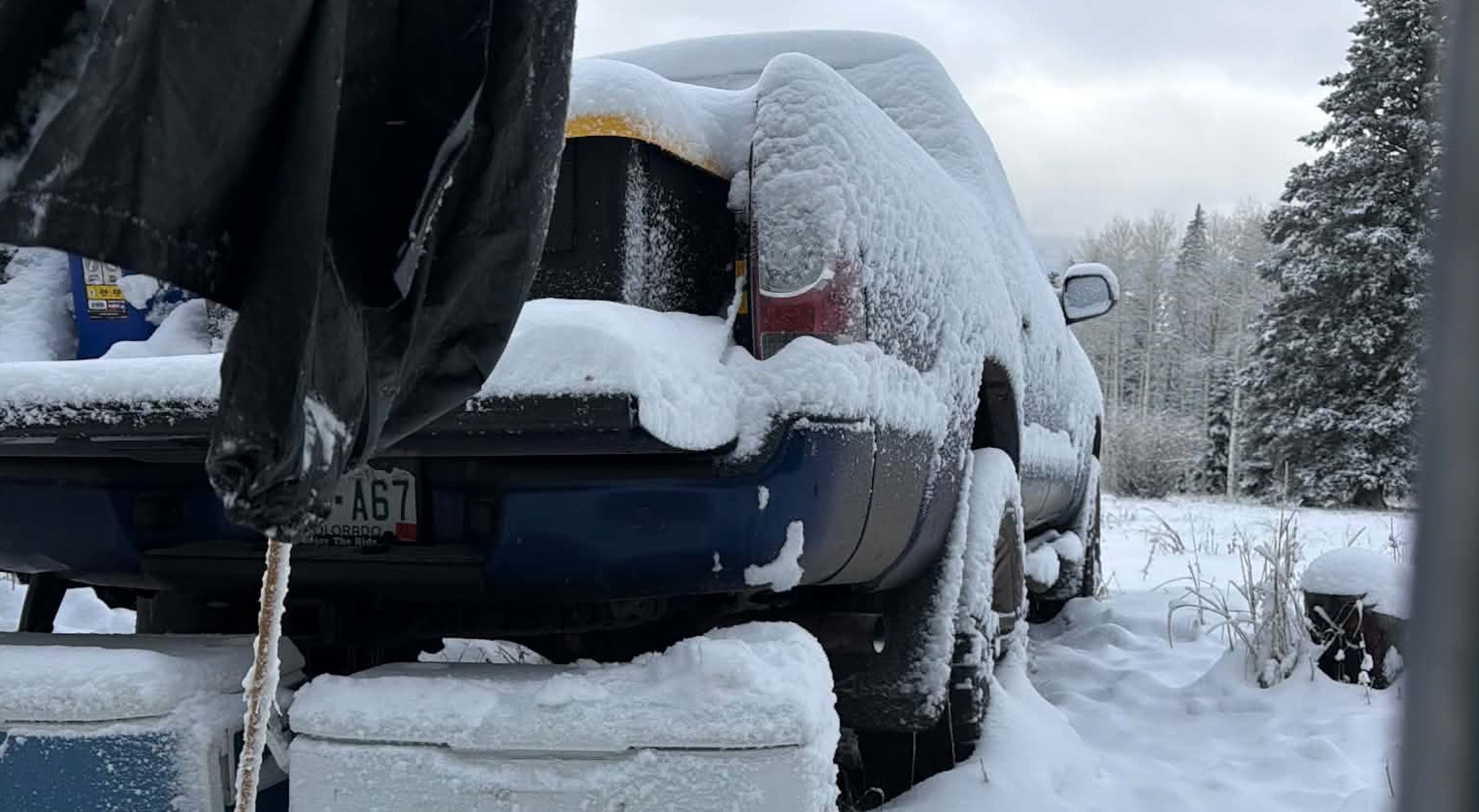 A pickup truck, storage containers, and a hanging tarp covered in fresh snow at a wooded hunting campsite in Colorado