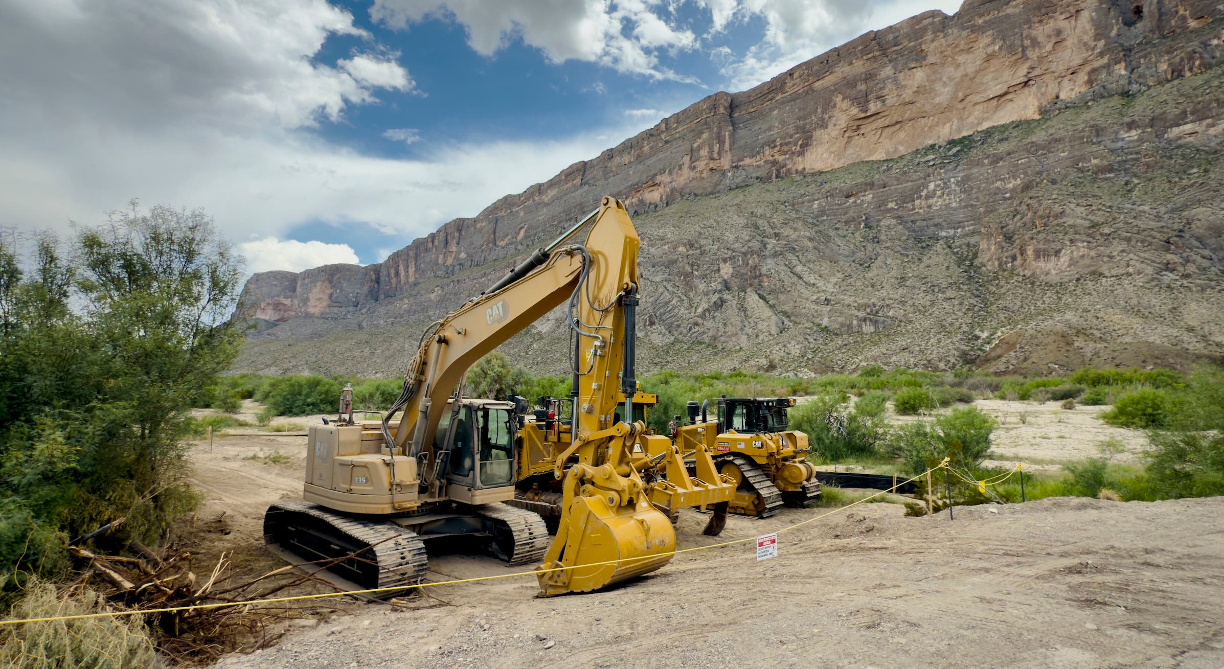 A yellow excavator and bulldozer sit on cleared dirt near Santa Elena Canyon in Big Bend National Park, with towering limestone cliffs and desert vegetation in the background.