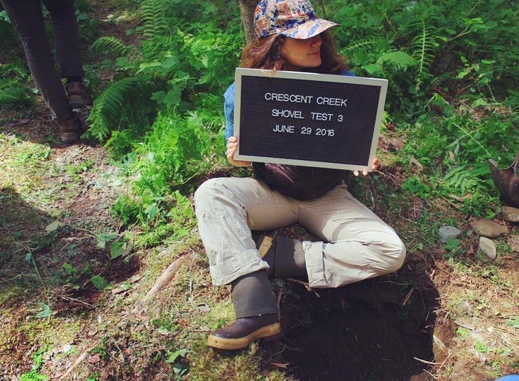 Author Madison Dapcevich sits above an excavation site holding a sign at Crescent Creek Campground