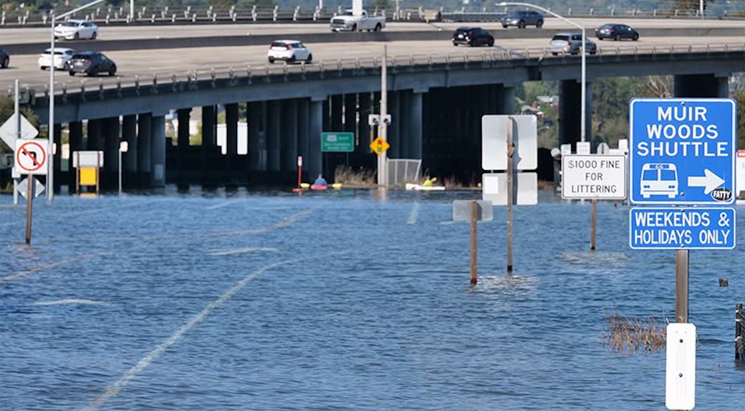 Kayakers paddle through a flooded roadway beneath a busy highway overpass in Marin County. Road signs, including a blue Muir Woods Shuttle sign, rise above the water, which covers the pavement and partially submerges lane markings.