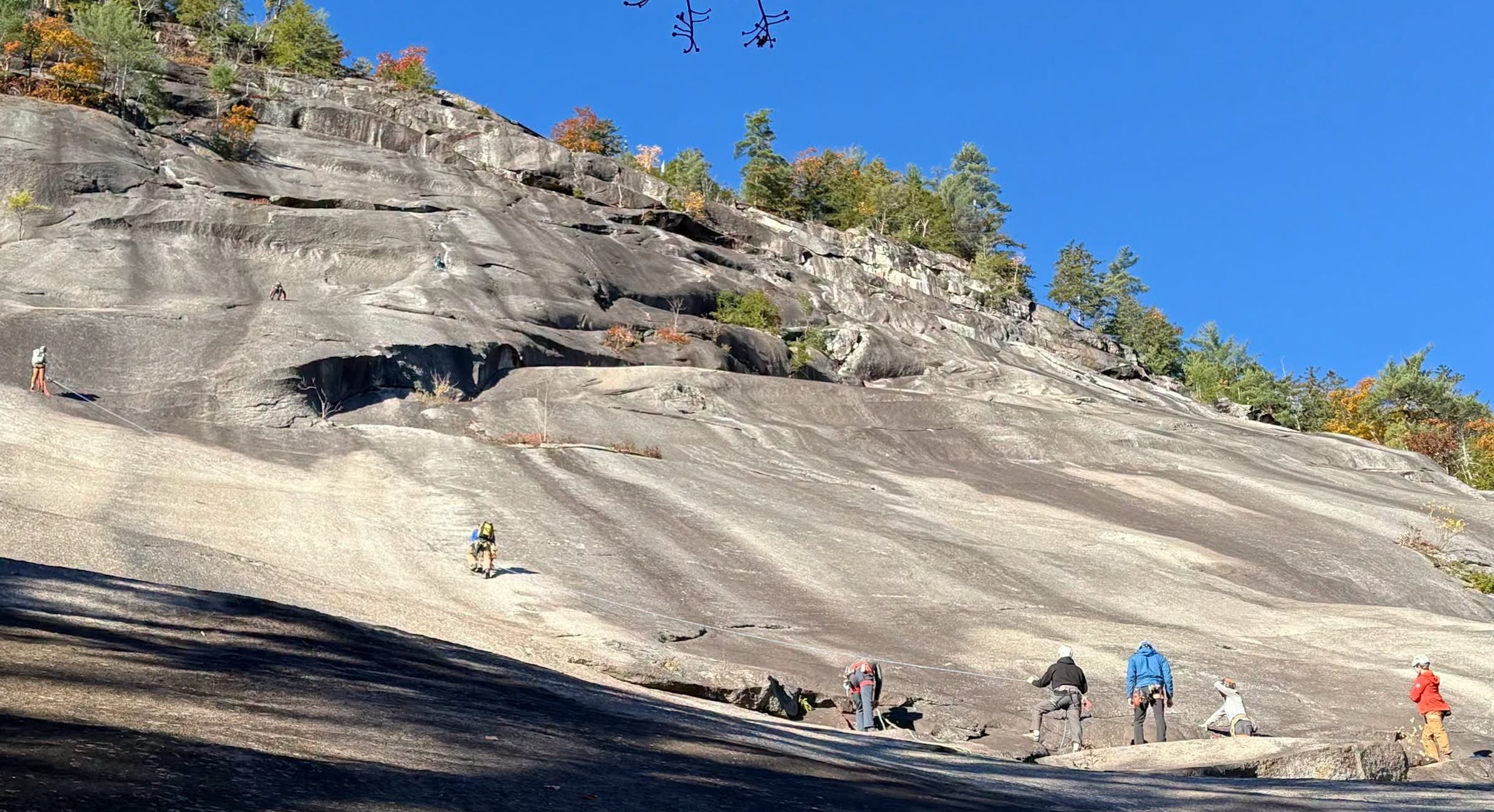 Climbers in helmets and harnesses handle ropes on Whitehorse Ledge’s sloping granite face, with other climbers higher on the rock and autumn trees above.